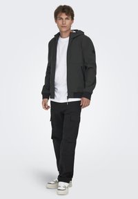 Zwarte ritssluitingjack met een capuchon, met zijzakken en ribgebreide manchetten, gedragen over een wit t-shirt en gecombineerd met zwarte cargobroek en sneakers.