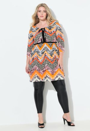 Eine Frau in einem gemusterten Kleid mit Zickzack- und Leopardenmotiven posiert vor einem schlichten Hintergrund und trägt schwarze Leggings und High Heels.