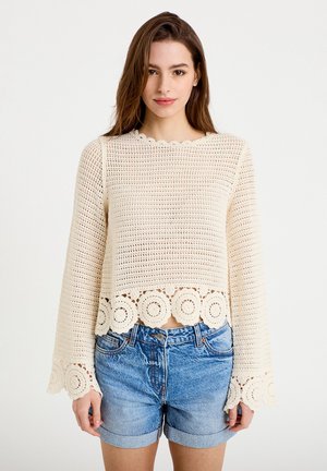 Giovane donna che indossa un top a maniche lunghe in crochet color crema con motivi circolari e pantaloncini di jeans blu risvoltati su uno sfondo semplice.