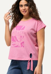 Růžové krátké tričko s květinovou grafikou a textem. Obsahuje větší uzel na lemu a detaily vyříznutého otvoru.