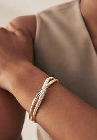 Guldtonet armbånd med sammenflettede bånd og en struktureret, funklende detalje. Glat overflade med et strømlinet design.