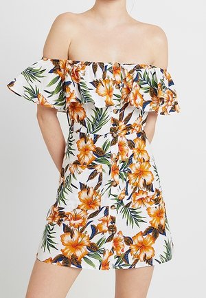 Mujer que lleva un vestido blanco de escote off-shoulder con estampado de flor de hibisco naranja y hojas verdes, detalle de botones en la parte delantera y escote con volantes.