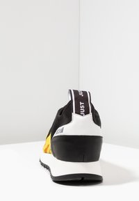 Zapatilla deportiva negra y amarilla con una parte superior texturizada, un contrafuerte negro sólido y una prominente lengüeta blanca con la inscripción "JUST".