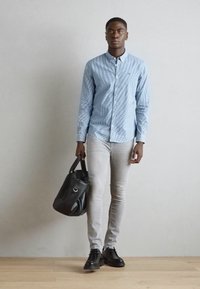 Light blue checkered shirt, gray slim-fit pants, black leather shoes. Holding a black tote bag, standing on a wooden floor against a plain wall.
