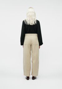 Asmuo su ilgu bangų plaukų plaukų, platininiu atspalviu, vilkintis juodą ilgą rankovę marškinėlį, smėlio spalvos plačiąsias kelnes ir tamsiai rudas batus, atsukęs nugarą.