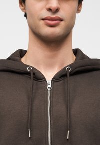 Sweat à capuche marron avec fermeture éclair, doté de cordons de serrage, fabriqué en tissu doux. Comprend une fermeture éclair argentée et deux accents métalliques sur les cordons.