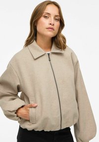 Bēšā, pārāk liela bomber jaka ar apakšējo rāvējslēdzēju, lielu apkakli un ribotām piedurknēm. Mīksts, teksturēts audums; ar sānu kabatām.