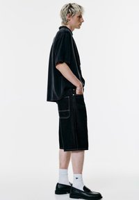 Ifjú ember rövid szőke hajjal, fekete, oversized rövid ujjú pólót, fekete térdig érő sortot, fehér zoknit és fekete loafert viselve, profilból.