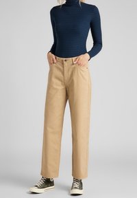 Beige ribbstickade byxor med raka ben och framfickor, tillsammans med en åtsittande mörkblå ribbstickad polotröja och gröna högskurna sneakers.