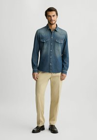 Camisa de denim con un degradado azul deslavado, dos bolsillos en el pecho y botones a presión. Se combina con pantalones beige claros y zapatos negros.