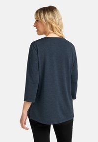 Blonde Frau, die seitwärts steht, trägt ein dunkelblaues Langarmshirt und schwarze Hose vor einem schlichten weißen Hintergrund.