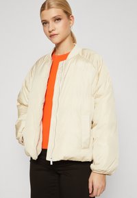 Pufferjakke i krem, med glidelås foran, ribbet krage og elastiske mansjetter, kombinert med en lys orange ribbet topp og svarte bukser.