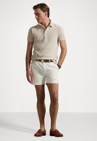 Beige polo-paita ja valkoiset sortsit, ruskea vyö, ruskeat loaferit. Polossa on pieni logo, sortsit ovat muotoonommeltuja sivutaskuilla.