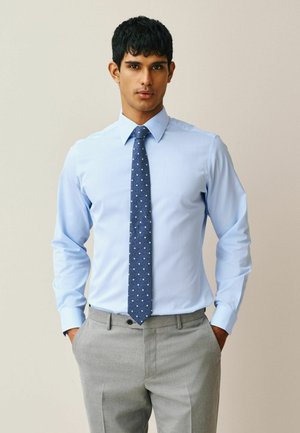 Young man wearing a light blue dress shirt, blue polka dot tie, and gray dress pants, standing with hands in pockets.