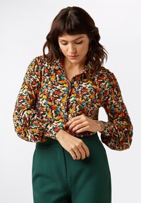 Multicolored floral blouse with a button front, long puff sleeves, and a collar. Paired with high-waisted green trousers.