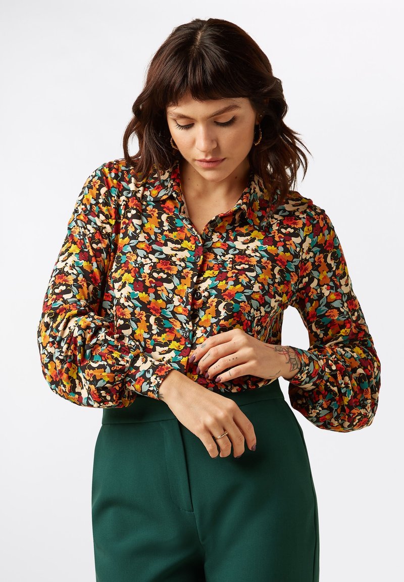 Multicolored floral blouse with a button front, long puff sleeves, and a collar. Paired with high-waisted green trousers.