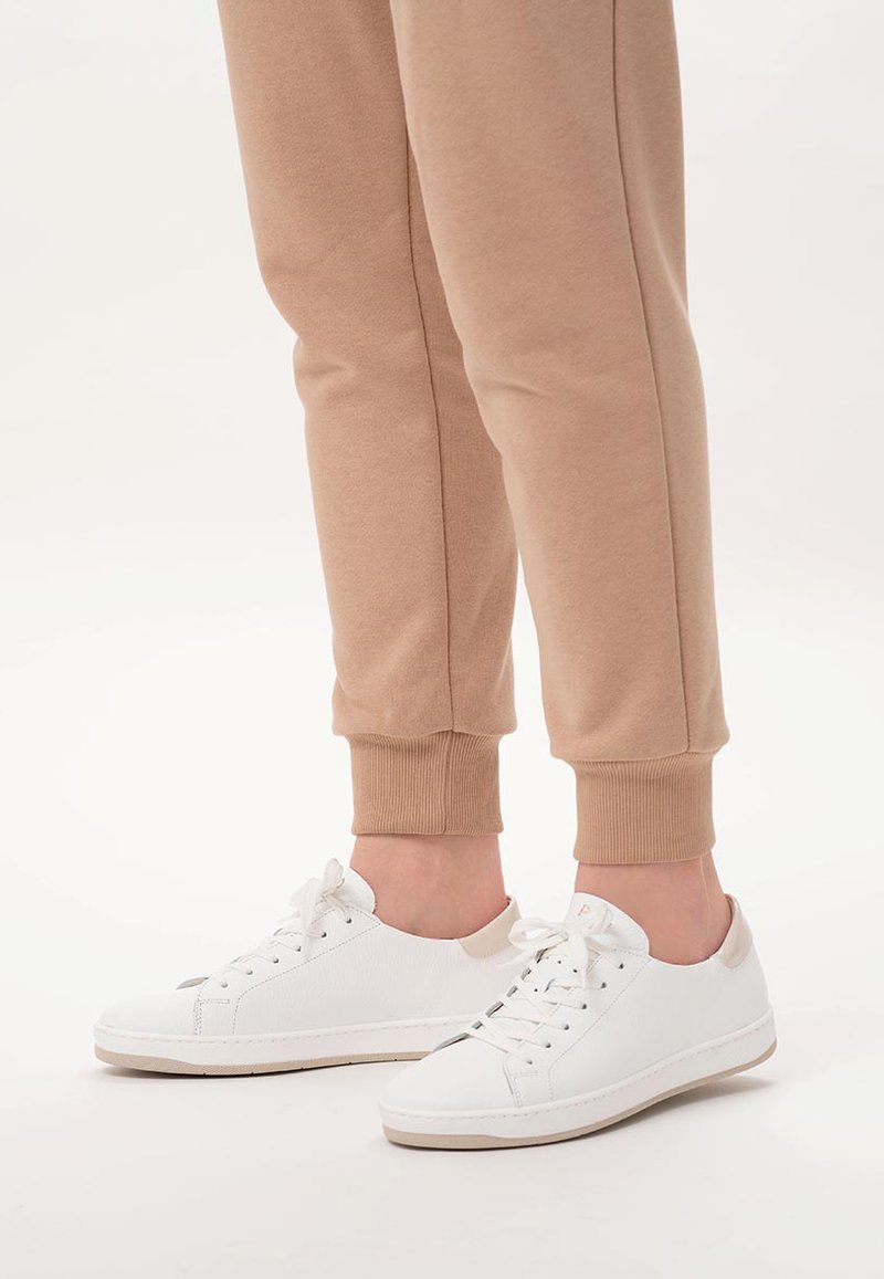 Białe sneakersy o gładkiej fakturze, z sznurowanym zapięciem i beżowym akcentem na pięcie. Połączone z beżowymi dresami z mankietami.
