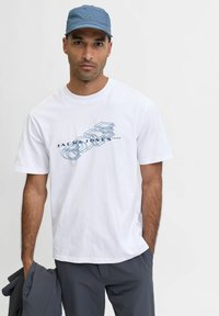 Bílé bavlněné tričko s modrým grafickým designem, krátké rukávy a volný střih, v kombinaci se šedými kalhotami a modrou čepicí.