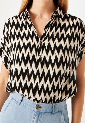 Nainen, jolla on musta-beigen siksak-kuosinen lyhythihainen paita, joka on tungettu vaaleansinisisiin korkeavyötäröisiin housuihin, joissa on nappi.