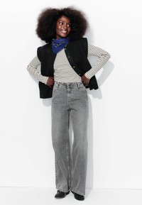 Veste noire par-dessus une chemise à manches longues rayée, associée à un jean gris taille haute à jambes larges et des chaussures noires à pois. Accent de bandana bleu.