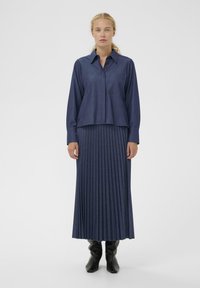 Koszula dżinsowa z kołnierzykiem i kieszenią, połączona z plisowaną, długością do podłogi spódnicą w ciemnoniebieskim kolorze. Noszona z czarnymi kozakami do kolan.