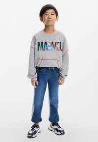 Šedá mikina s multibarevným grafikou "MARVEL", světle modré džíny se postranním pruhem a černo-bílé tenisky s barevnými akcenty.