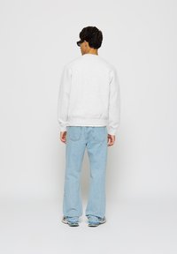 Man with short dark hair wearing black glasses, light gray sweatshirt, light blue jeans, and sneakers, standing facing a white background.