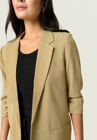 Beige színű, szabott blézer, hegyes hajtókával, felhajtott ujjal és egyetlen elülső zsebbel, egy fekete bordás felső felett viselve. Sima szövettextúra.