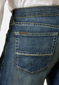 Pantalones vaqueros de mezclilla azul oscuro con un acabado ligeramente texturizado, costuras amarillas y una pequeña placa de logotipo marrón en el bolsillo trasero.