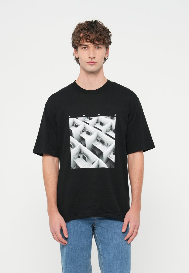 Νεαρός με σγουρά μαλλιά που φοράει μαύρο T-shirt με ασπρόμαυρο σχέδιο γραφείου σε θήκη και το κείμενο «HUGO», συνδυασμένο με μπλε τζιν.