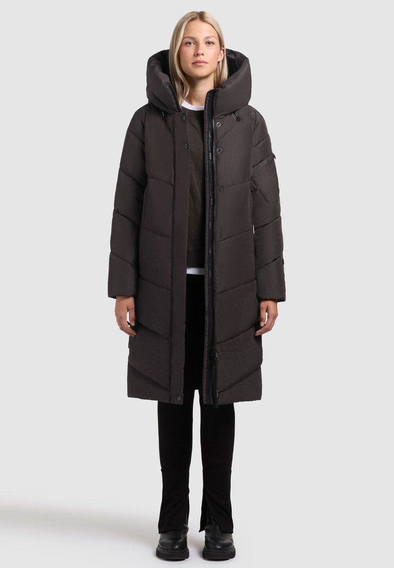 Kvinna iklädd en lång svart pufferjacka med stor huva, svarta byxor och svarta stövlar, stående mot en ljusgrå bakgrund.