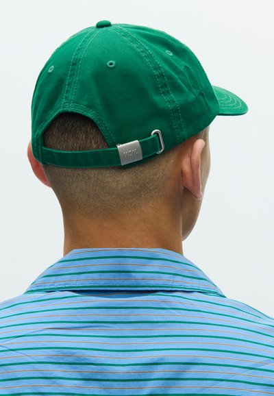 Πράσινο βαμβακερό καπέλο με κυρτή γείσο, ρυθμιζόμενο λουράκι με μεταλλικό στοιχείο. Ριγέ μπλε πουκάμισο εμφανίζεται στο φόντο.