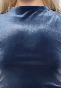 Laivastonsininen samettitoppi, jossa on sileä tekstuuri ja korkea, pyöreä kaula-aukko. Kangas hehkuu hienovaraisesti ja siinä on kevyitä ryppyjä.