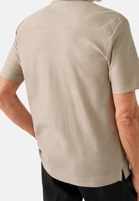 Camicetta beige a maniche corte con tessuto a trama, dotata di colletto classico e spacchi laterali, vista da dietro.