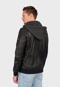 Sort læderjakke med grå hætte, ribbede manchetter og kant. Bagindsigt viser sømdetaljer og et logo-patch på venstre ærme.