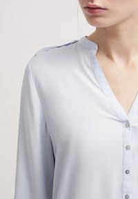 Ljusblå blus med V-ringning, knappstängning framtill och mjukt, slätt tyg. Har kontrasterande sömmar längs kragen och axlarna.