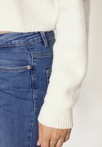 Pull en maille crème avec une texture côtelée, associé à une jupe en jean bleu dotée de cinq poches et de détails en métal argenté.