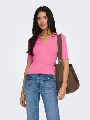 Kvinne med langt mørkt hår iført en rosa ribbestrikket kortermet topp og blå jeans, bærer en stor brun skulderveske.