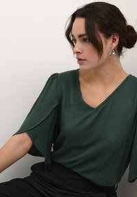 Žalia tekstūruota bluzonė su V formos iškirpte ir laisvais rankovėmis, derinama su juodomis aukštai iškelta juosmens kelnėmis. Pasižymi subtiliais deimantų raštais ir minkštu kritimu.