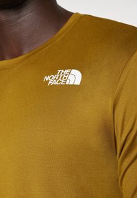 Camisa de manga longa amarelo-escura com decote arredondado. Apresenta o logo branco "The North Face" na zona superior esquerda do peito. Textura suave.