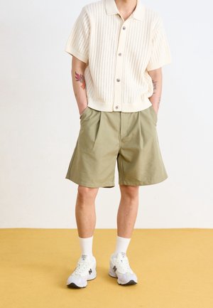 Cremefarbenes Strick-T-Shirt mit kurzen Ärmeln und Knöpfen, kombiniert mit olivgrünen plissierten Shorts. Weiße Sneaker mit grauen Akzenten und gerippte Kurzsocken.