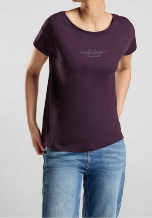 Kobieta w ciemnofioletowej koszulce z napisem "mindfulness Let's grow together" i jasnoniebieskich dżinsach, stojąca na jednolitym tle.