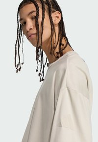 Svetlo béžová mikina s dlhým rukávom vyrobená z jemnej tkaniny, s klasickým výstrihom a minimalistickým logom na hornom rukáve.