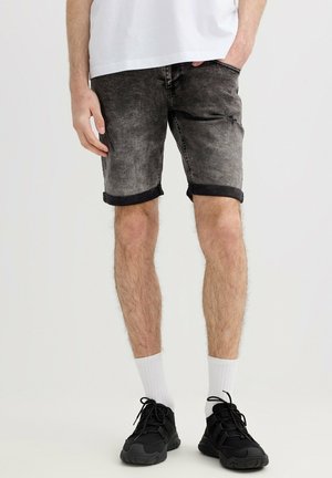 Hombre con pantalones cortos de mezclilla negra desgastada, calcetines blancos, zapatillas negras y una camiseta blanca, de pie con una mano en el bolsillo.