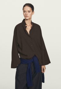 Bruine oversized button-up shirt met een kraag, gemaakt van een gestructureerde stof, gedragen over donkerblauwe broek met een gestrikt ceintuur. Eenvoudige designelementen.