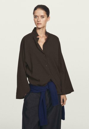 Camicia oversized marrone con bottoni e colletto, realizzata in un tessuto texturizzato, indossata sopra pantaloni blu navy con una cintura annodata. Elementi di design semplici.