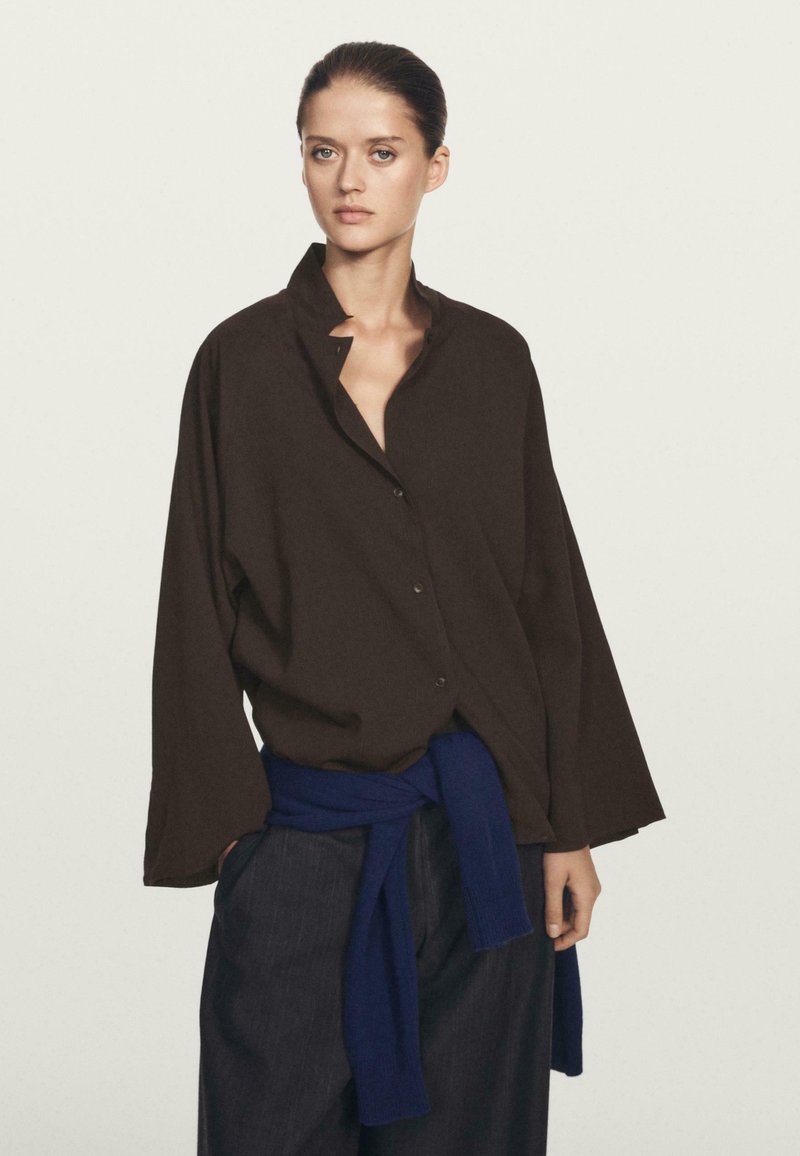 Camicia oversized marrone con bottoni e colletto, realizzata in un tessuto texturizzato, indossata sopra pantaloni blu navy con una cintura annodata. Elementi di design semplici.