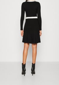 Vestido de punto acanalado negro con mangas largas y falda acampanada. Presenta un detalle de cinturilla blanca. La modelo lleva botas negras hasta el tobillo.