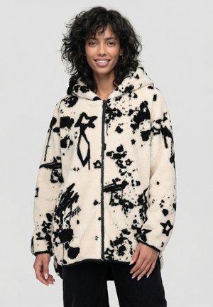 Frau mit lockigem schwarzen Haar, die einen weißen Fleece-Kapuzenpullover mit abstrakten schwarzen Spritz- und Sternmustern trägt, lächelt und vor einem schlichten Hintergrund steht.