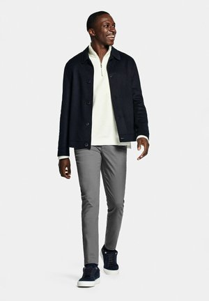 Smiling man walking, wearing a black jacket, white half-zip sweater, gray pants, and dark blue sneakers with white soles on a white background.